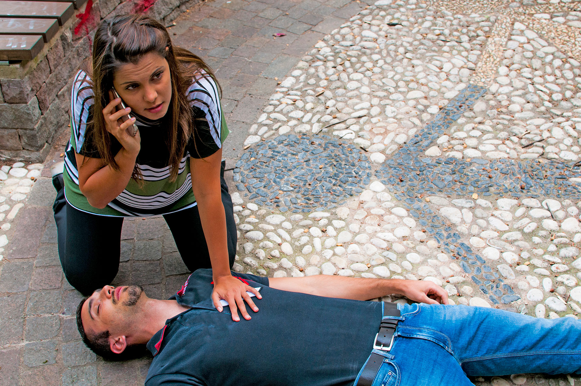 A person lies on the ground with his eyes closed while another person crouches next to him and talks on the phone with a worried look