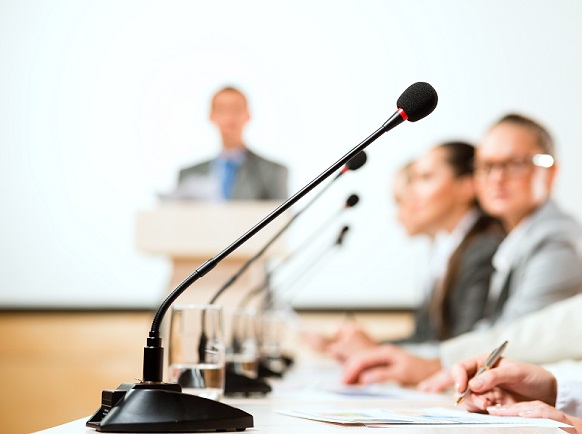 People in a row sit behind microphones that are on a table, with another person standing at a lectern in the background